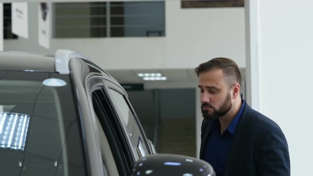 Stylish guy is choosing new automobile walking around luxury car and touching it in car dealership