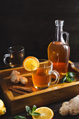 Homemade Kombucha in a glass decanter and glasses on a wooden tray with ginger, mint and lemon on a dark background. This is a healthy natural fermented sweet and sour drink originally from China