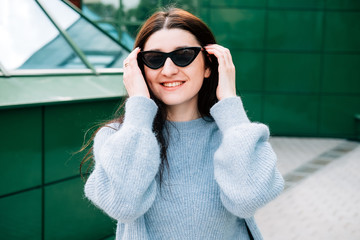 Outdoors portrait of beautiful young brunette girl smiling, close up. Teenager hipster girl with sunglasses wearing trendy outfit posing in city street. Urban style concept, green background.