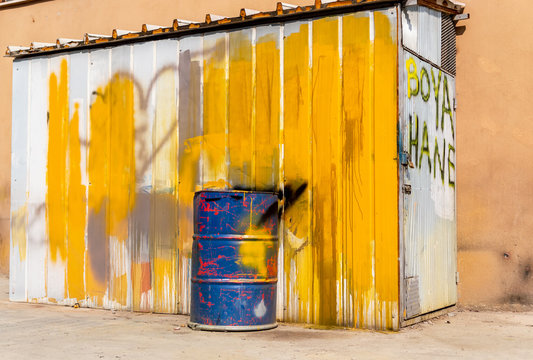 Blue metal drum in front of a shed painted in white and yellow. Text on the door: Dye house