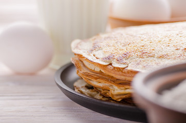 Stack of French crepes in frying pan on wooden kitchen table with milk eggs and flour aside