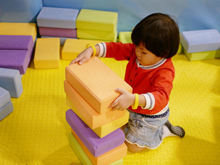 Little Asian baby girl stacking up foam building bricks / blocks at an indoor playground - playing foam blocks helps develop children's fine motor and logical thinking skills