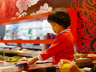 Little Asian baby girl enjoys picking raw ingredients, for hotpot cooking, serving on a moving conveyor belt