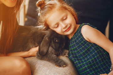 young and stylish mother with long hair and a green dress playing with her little cute daughter and little black rabbit at home