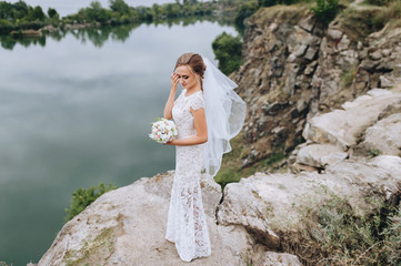 A young smiling and beautiful bride in a white dress is standing against a background of rocks,...