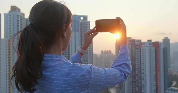 Woman enjoy the view of the city and take picture on mobile phone under sunset view