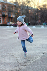 beautiful little girl in a hat, coat and jeans jumps, flies and has fun on the background of the cityscape in the light of sunset.
