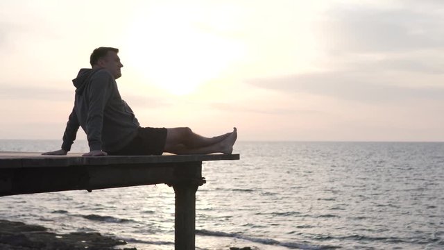 A man watches the sunset in the sea. He sits on a wooden bridge over a cliff. A tourist on vacation watches a beautiful sunset in the ocean. Spring in Cyprus