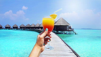 A glass of orange juice in traveler's hand with sea and wooden bungalow background for summer holiday concept.