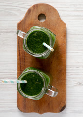 Green smoothie in glass jars on a rustic wooden board over white wooden surface, top view. Flat lay, overhead, from above.