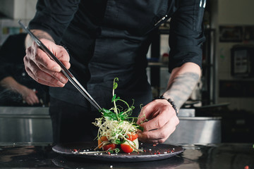 Chef finishing healthy salad on a black plate with tweezers. almost ready to serve it on a table
