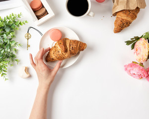woman's hand holding croissant.  White table's surface with macarons, coffee, and flowers. Flat lay with french style breakfast, copy space for your text
