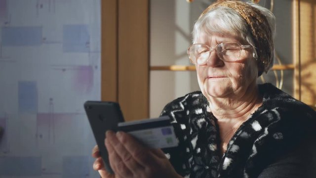 An old woman using a smartphone and a credit card examines payment documents. Social inequality