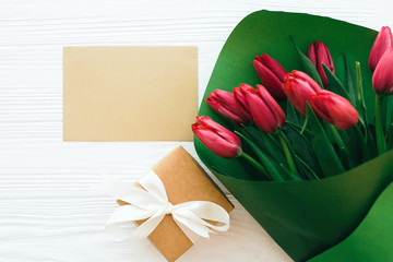 Beautiful red tulips bouquet in green paper,gift box and craft card with space for text on white wooden background flat lay. Happy mothers day. Pink tulips on white wood. Greeting card template