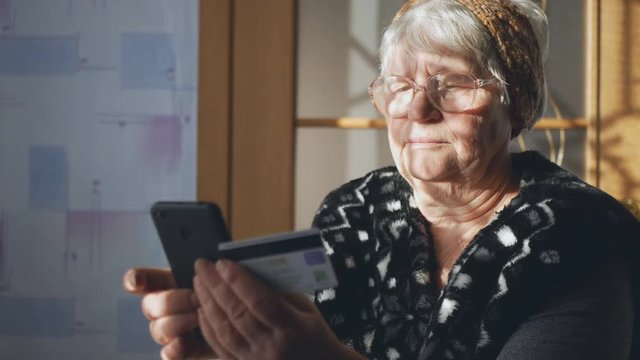 An old woman using a smartphone and a credit card examines payment documents. Social inequality