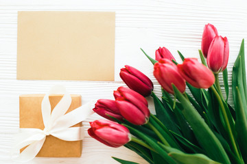 Beautiful red tulips with gift box and craft card with space for text on white wooden background, flat lay. Happy Mothers day. Pink tulips on white wood. Greeting card template. Valentines day