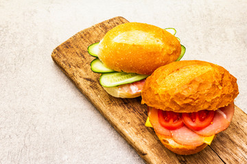 Homemade sandwich in bun. Salami, dried pork, tomato, cucumber, cheese ingredients. On wooden board and stone background.