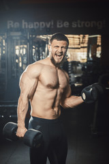 Fototapeta na wymiar Good looking young man bodybuilder lifting dumbbells working on his biceps screaming shouting in front of the mirror at the gym. Athlete holding dumbbells showing straining veins on hands bubble guts