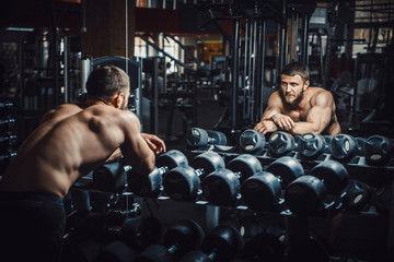 tired guy bodybuilder bowed his head in the gym because lack of strength and energy. athletic man waiting and resting before lifting dumbbells in front of the mirror. athlete needs rest and motivation