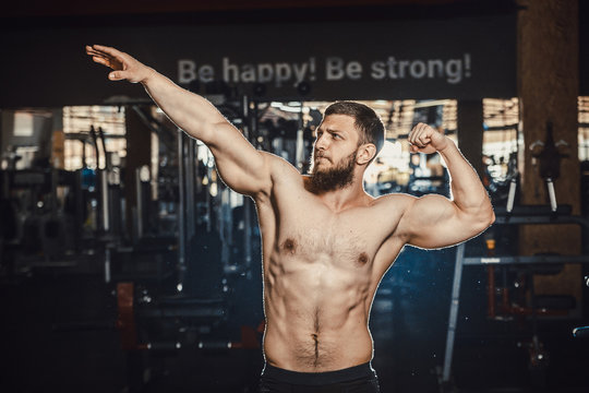 Good looking young man bodybuilder posing in front of the mirror shows big biceps at the gym darkened slogan background. Athlete showing straining veins on hands bubble guts. arnold pose