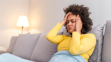 Young woman sitting on a living room couch, covered with a blanket, holding her head, having a splitting headache and suffering enormous pain