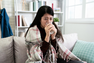 Fototapeta na wymiar japanese beautiful woman wrapped in warm blanket holding drinking glass of water freezing sitting on sofa at home. sick asian lady cover by quilt on couch near window looks upset frustrated.
