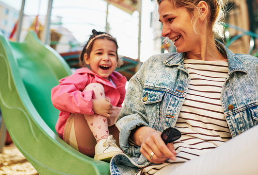 Candid image of happy young woman laughing with her little cute daughter spending time together outdoors on slide at playground. Mother with her toddler girl feeling happy, loving each other outside.