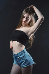 Young beautiful, slender, passionate brown-haired woman with bright red lips and long hair in a black top and denim shorts on a dark background