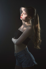 Young beautiful, slender, passionate brown-haired woman with bright red lips and long hair in a black top and denim shorts on a dark background