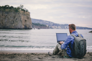 man, guy the photographer working with a laptop on the beach, backpack, concept of find information at travel to foregin country, communication, mountains on background