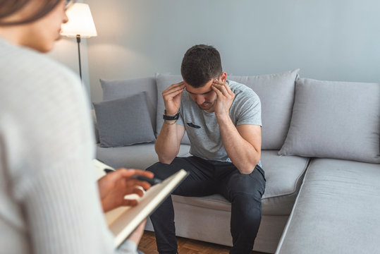 Handsome young man is sitting on couch and talking to the psychologist while doctor is making notes. Young man consulting with medical doctor or psychologist, complaining about health