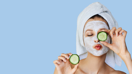 Beautiful young woman with facial mask on her face holding slices of cucumber. Skin care and treatment, spa, natural beauty and cosmetology concept. - obrazy, fototapety, plakaty