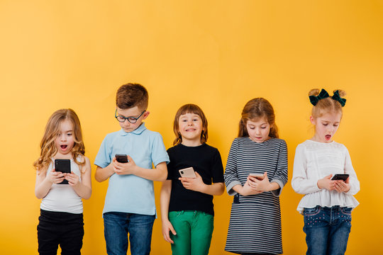 5 beautiful children use smartphone, surprised, happy, little boy in glasses and 4 little girls are happy, different emotions,, isolated yellow background, copy space