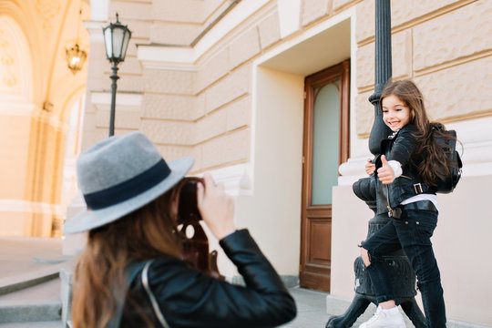 Lovely schoolgirl with black backpack gladly posing on the street leaning to the iron tube and showing thumb up. Elegant woman in vintage hat making photo of little daughter during walk around city.