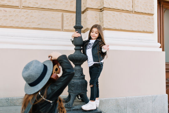 Cute little girl with funny face expression standing near the iron pillar, posing in front of mother in trendy felt hat. Young woman with professional camera making photoshoot for daughter.