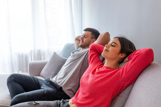 Couple relaxing on a sofa at home and looking at TV at the living room. Side view of a happy couple breathing and resting lying in a couch at home with a window in the background