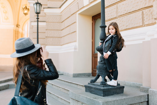Cute brunette girl wearing white sneakers and denim pants holding by pillar, while mother taking picture standing in front of her. Elegant young woman carrying leather bag and camera making photo.