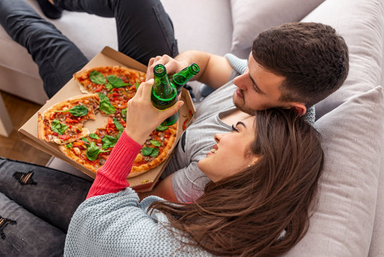 Cheerful couple relaxing together on couch and watching movie at home.They are laughing and eating pizza and having a great time. Happy couple with pizza clinking by beer bottle