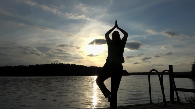 eine Frau steht bei Sonnenuntergang am See und macht Yoga