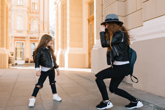 Cute little girl in black pants with holes on knees posing on the street, while her mother taking photo. Graceful young woman with backpack and camera making picture of joyful daughter in sunny day.