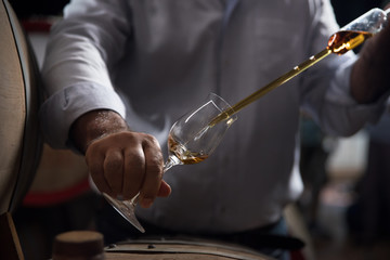 Man, pouring cognac from the barrel into glass in old rustic underground wine cellar with rows of big oak barrels. Famous wine and brandy industrial destination - obrazy, fototapety, plakaty