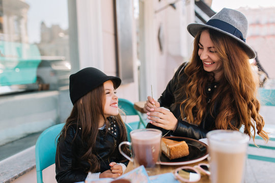 Pretty young mother and her cute daughter delicious breakfast with coffee and dessert in a cafe on the city background. Styling family, true emotions, good day.