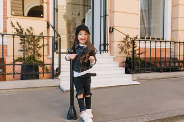 Full-length portrait of excited kid in grunge outfit funny screaming outside in front of shop. Playful little girl with long brown hair wearing leather clothes fooling around on the street