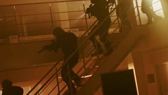 Masked Squad of Armed SWAT Police Officers Run Down the Stairs from a Second Floor in a Dark Office Building. Soldiers with Rifles, Flashlights Move Forwards and Cover Surroundings. Warm Color Grading