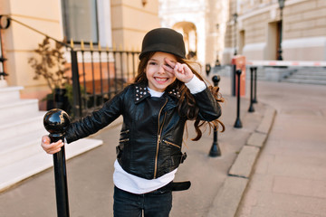 Portrait of laughing little girl in trendy black jacket and white shirt waiting friends on the street. Cute long-haired kid with amazing smile posing outside on city background after lessons