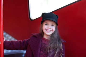 Fototapeten Kleines fröhliches Mädchen in der Nähe des roten englischen Busses in einem schönen Mantel und Hut. Kleines fröhliches Mädchen in der Nähe des roten englischen Busses in einem schönen Mantel und einem Hut. Reise des Kindes. Schulbus. Roter Londoner Bus. © stock_studio