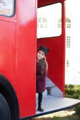 Schilderijen op glas Little cheerful girl near the red English bus in a beautiful coat and a hat. Child's journey. School bus.  London red bus. Spring.  With the International Women's Day. Since March 8! © stock_studio