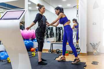 Fototapeta na wymiar Young woman fitness instructor works with woman wearing ems equipment as she exercises in a studio. Smiling satisfied female coach training woman