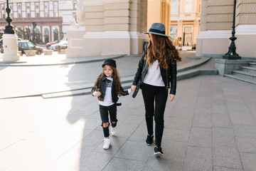 Slim stylish woman in black jeans holding hand of her cute daughter in leather jacket, walking down the street. Portrait of young mother in felt hat and cheerful long-haired girl having fun in city