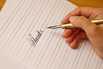 female hand writes with the inky pen the word thanks on a white paper sheet with stripes. stationery on desk close up top view. spelling lessons and caligraphy exercises. Template, layout, background
